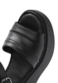 Sandale en cuir noir avec des larges brides rembourrées, une finition texturée et une semelle plateforme. Des détails de couture le long des bords sont visibles.