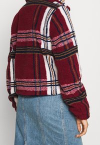 Veste en polaire à carreaux rouges et blancs avec une surface texturée et des rayures bleu marine, marron et rose. Associée à une jupe en denim bleu clair.