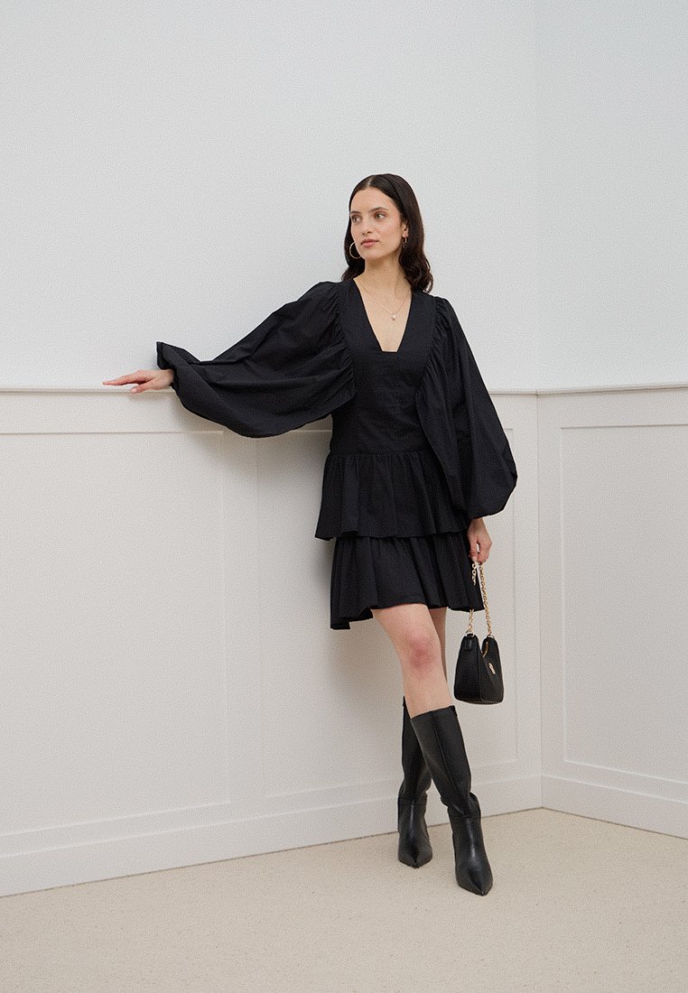 Schwarzes Mini-Kleid mit voluminösen langen Ärmeln, geschichtetem Rüschensaum und tiefem V-Ausschnitt. Kombiniert mit kniehohen schwarzen Stiefeln und einer kleinen Handtasche.