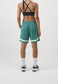 Nainen, jolla on pitkä letitys, pukeutuneena mustaan urheiluliivimekkoon, vihreisiin urheilushortseihin, joissa on valkoiset raidat, valkoisiin Nike-sukkiin ja mustiin sekä valkoisiin lenkkareihin, seisoo.