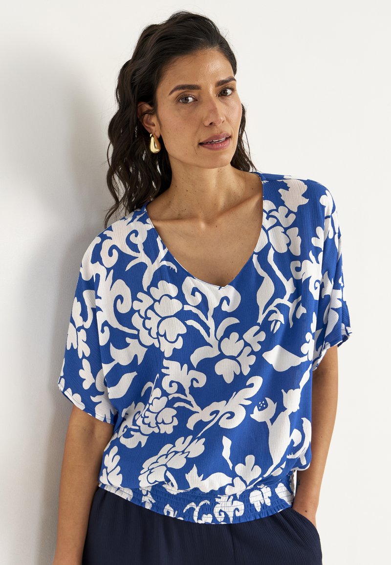 Blå blomsterbluse lavet af tekstureret stof. Har V-hals, løs pasform og hvide blomster mønstre. Rynket nederste kant med elastik.