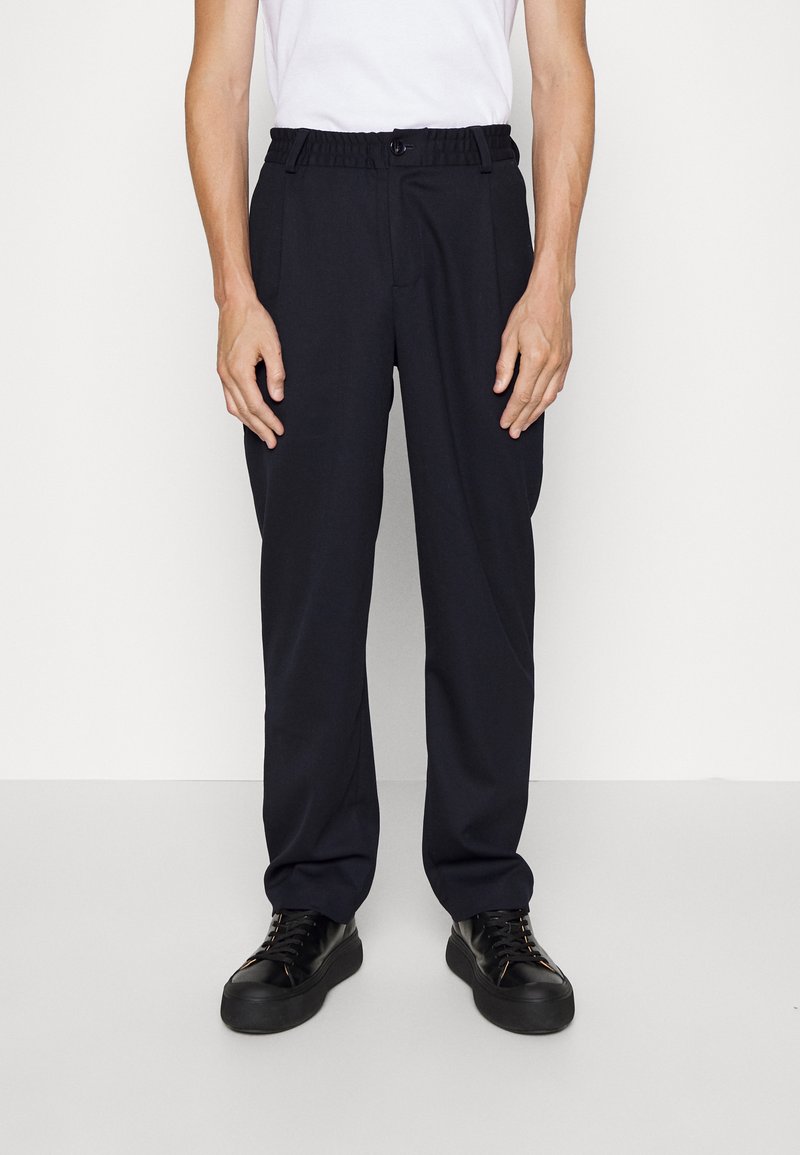 Schwarze Hose mit elastischem Bund, zwei Seitentaschen, geradem Schnitt und glatter Textur. Getragen mit schwarzen Turnschuhen vor einem hellen Hintergrund.