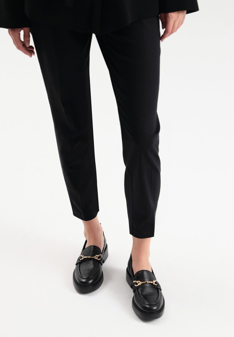 Czarne skórzane mokasyny z teksturowanym wierzchem, ze złotymi detalami, w połączeniu z dopasowanymi czarnymi spodniami, ukazującymi smukłą sylwetkę.