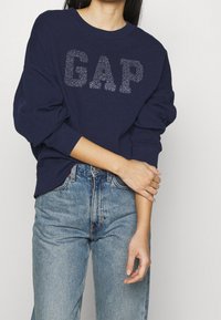 Námořnicky modrá mikina s výrazným stříbrným logem "GAP", volný střih, kulatý výstřih a dlouhé rukávy, doplněná o modré džíny s vysokým pasem.