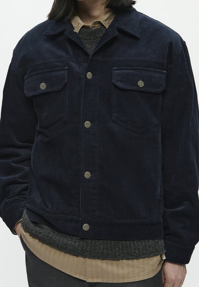 Námořnická kordáčová bunda se dvěma náprsními kapsami, stříbrnými patentkami, límcem a strukturovaným povrchem. Nositá přes šedé a béžové základní oděvy.