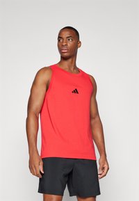 Rde majica brez rokavov za šport z črnim Adidasovim logotipom na prsih. Tkanina izgleda gladka in lahka, v kombinaciji s črnimi kratkimi hlačami.