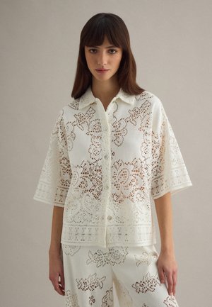 Mujer joven con una camisa blanca de encaje abotonada y pantalones de encaje a juego, posando contra un fondo neutro y liso.