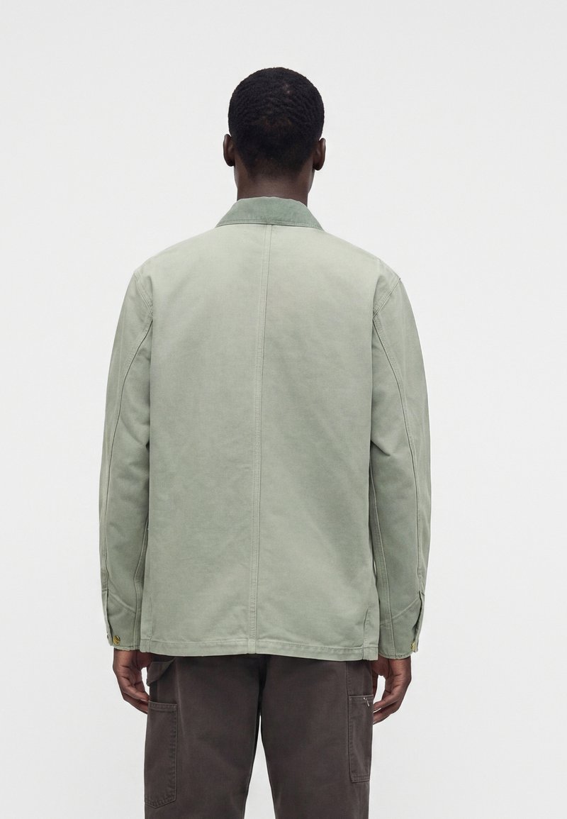 Mann von hinten gesehen, trägt eine hellgrüne Jacke mit sichtbaren Nahtdetails und dunkle Hosen vor einem einfarbigen hellen Hintergrund.
