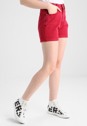 Persoon draagt rode denim shorts, witte hoge sneakers met zwarte letters en een wit topje met één hand in de zak.