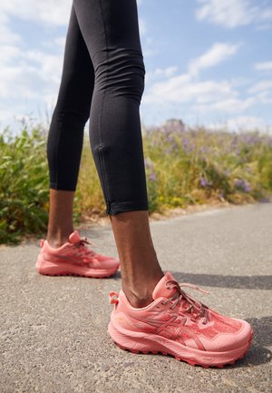 Close-up of feet wearing pink running shoes and black leggings standing on a paved path with green plants and sky in the background.