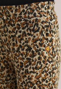 Tessuto con stampa leopardata in oro, marrone e nero, caratterizzato da una cintura elastica e un piccolo tag con logo arancione sulla cucitura laterale.