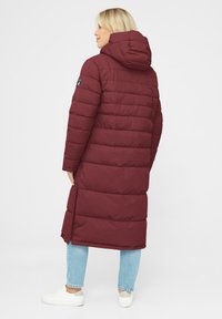Burgundy puffer coat with a hood, long design, horizontal stitching, side slits, and a logo patch on the left sleeve. White sneakers and blue jeans.