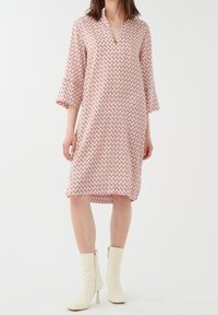 Robe imprimée avec un col en V, des manches cloche trois-quarts, de couleur crème clair avec un motif géométrique rouge et vert. Coupe au genou.