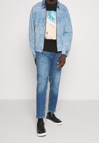 Chaqueta de denim azul claro sobre una camiseta gráfica negra con una escena de playa, combinada con jeans azul medio y zapatillas negras.