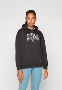 Felpa nera con cappuccio con stampa "PUMA sports club", tasca frontale e polsini a coste, abbinata a leggings azzurri chiari.