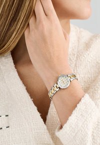 Hõbeda- ja kuldtooniga roostevabast terasest kell ümmarguse näoga, lillemustriga valgel taustal, kantud randmel, millel on tekstureeritud valge varrukas.