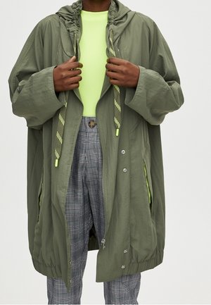 Personne vêtue d'une veste à capuche oversize vert olive par-dessus un haut vert fluo et un pantalon à carreaux gris, tenant les bords de la veste ouverts.