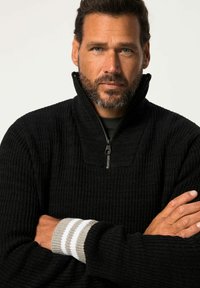 Maglione nero a costine con collare alto e chiusura a mezza zip. I polsini presentano dettagli a righe grigio chiaro e bianche.