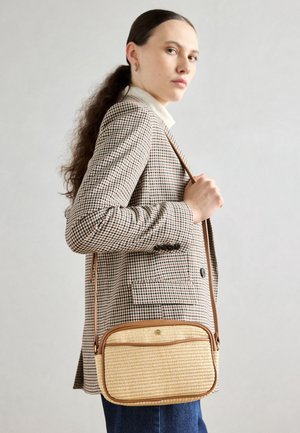 Tkaná slámová crossbody taška s hnědými koženými detaily, obdélníkového tvaru, s horním zipovým uzávěrem a odnímatelným popruhem, nošená na kostkovaném blejzru.