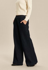 Brede donkerdenim broek met zijzakken, voorzien van een geraffineerde tailleband. Gekomplementeerd met een lichtgekleurde gebreide top.