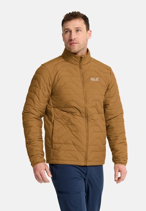 Man die een bruine doorgestikte jas met een voorrits en blauwe broek draagt, staat met een neutrale uitdrukking en kijkt iets omhoog.