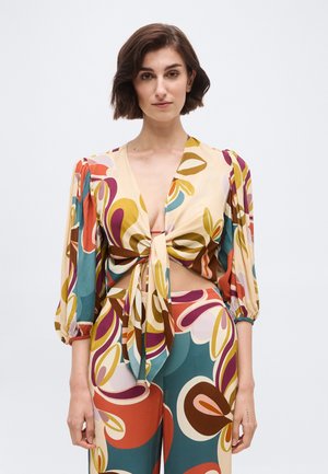 Femme aux cheveux courts bruns portant un chemisier noué à l'avant avec un motif floral multicolore, accompagné d'un pantalon taille haute assorti, sur un fond uni.
