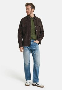 Jachetă din suede maro, cu două buzunare pe piept, tricou verde cu guler rotund dedesubt, blugi bleu deschis și adidași albi cu accente negre.