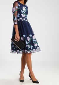 Marineblauwe jurk met bloemenborduurwerk in pastelkleuren, doorschijnende driekwartmouwen, aansluitend lijfje en uitlopende rok. Zwarte clutch en hakken.