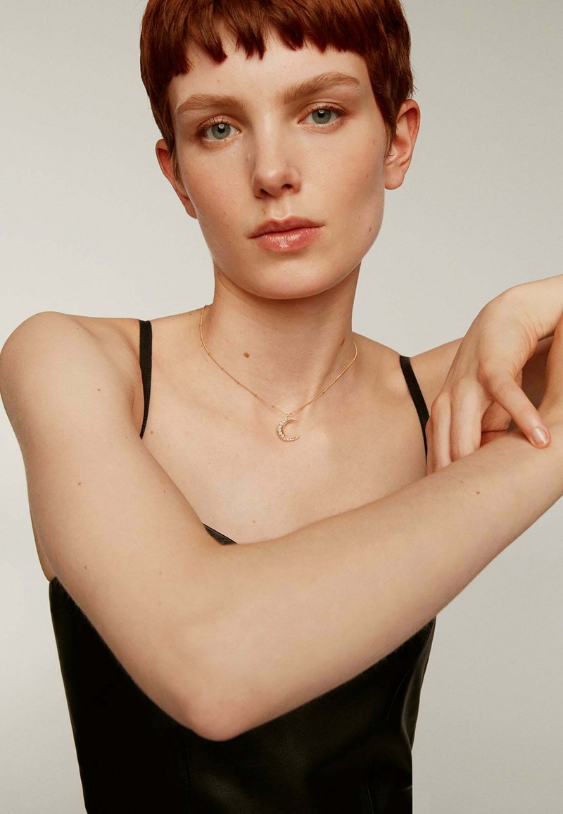 Collar de oro con un colgante en forma de luna creciente adornado con pequeñas gemas, llevado sobre una camiseta negra ajustada. El tono de piel es claro y tiene un maquillaje mínimo.