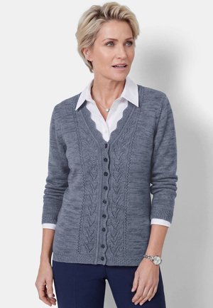 Femme portant un cardigan en tricot bleu boutonné sur une chemise blanche à col et un pantalon foncé, debout devant un fond clair uni.