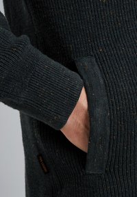Gebreide trui in donkergroen met een gestructureerd ribpatroon, uitgerust met een voorkwam en een label. Het ontwerp is praktisch en casual.