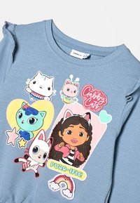 Gaisma zila garām piedurknēm krekls ar multiplikācijas varoņu attēliem un krāsainiem rakstiem, ar volānu rotājumiem pie pleciem.