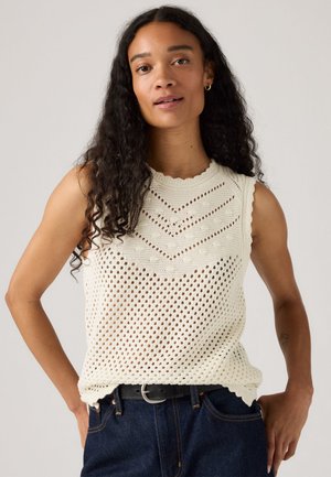 Woman with long curly hair wearing a sleeveless cream knit top with a chevron and openwork pattern, dark jeans, and a black belt.