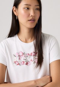 Άσπρο βαμβακερό t-shirt με λογότυπο Levi's σε λουλουδάτο σχέδιο σε ροζ και γκρι, με κλασική στρογγυλή λαιμόκοψη και κοντά μανίκια.