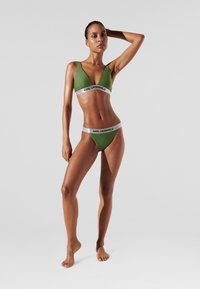 Žalias bikinis, kurį sudaro trikampis braletė ir žemos talijos kelnaitės, akcentuotos sidabriniu diržu su užrašu "KARL LAGERFELD."
