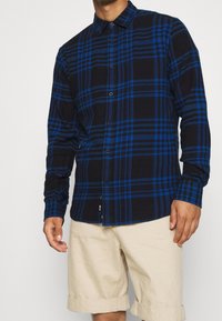 Camicia a quadri nera e blu con bottoni, maniche lunghe, colletto classico e tessuto liscio. Abbinata a pantaloncini di colore chiaro.