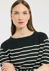 Frau mit glatten braunen Haaren, die einen schwarz-weißen gestreiften Pullover trägt, mit verschränkten Armen direkt in die Kamera schaut und einen neutralen Gesichtsausdruck hat.