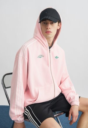 Giovane seduto su una sedia di metallo che indossa una felpa con cappuccio rosa Adidas con zip e cappuccio alzato, berretto nero e pantaloncini neri Adidas con strisce bianche.