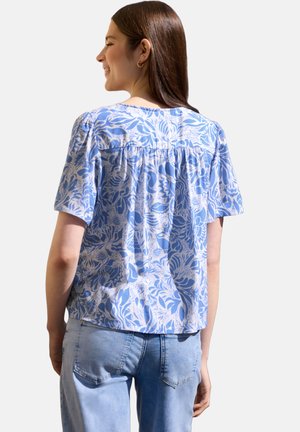 Kobieta ubrana w niebiesko-białą kwiecistą bluzkę z krótkim rękawem i jasnoniebieskie jeansy, zwrócona tyłem z delikatnym uśmiechem.