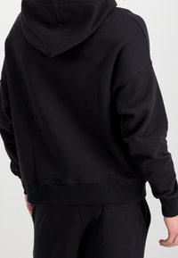 Felpa nera con cappuccio regolabile, vestibilità ampia e polsini a coste. Il materiale appare morbido e texturizzato, progettato per il comfort.