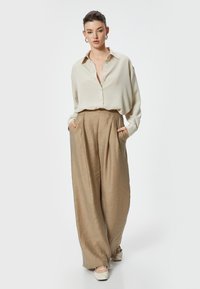 Camicetta beige a maniche lunghe con vestibilità rilassata e dettagli in bottoni, abbinata a pantaloni khaki a gamba larga con vita alta e tessuto testurizzato, e scarpe ballerine chiare.