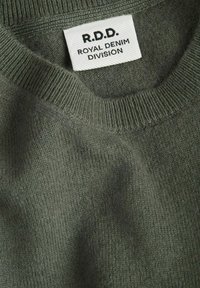 Gros plan sur l'encolure d'un pull en maille vert olive avec une étiquette blanche indiquant "R.D.D. Royal Denim Division".