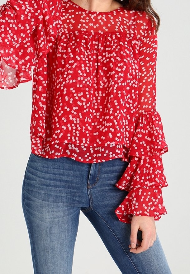 Blouse rouge transparent à pois blancs, manches évasées à volants et coupe décontractée. Associée à un jean bleu clair pour le contraste.