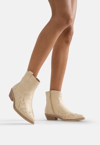 Botas de cuero crema hasta el tobillo con costuras decorativas, cremallera lateral, tacón bajo y apilado, y diseño de punta afilada. Acabado texturizado y estilo western.