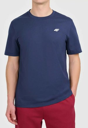 Mężczyzna w granatowej koszulce z krótkim rękawem z logo 4F i czerwonych spodniach, stoi z jedną ręką w kieszeni na jednolitym tle.
