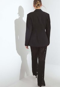 Svart skräddarsydd kavaj med skarpa slag kombinerad med genomskinliga svarta spetsshorts, vilket skapar en lager-på-lager textureffekt. Skugga syns på väggen.