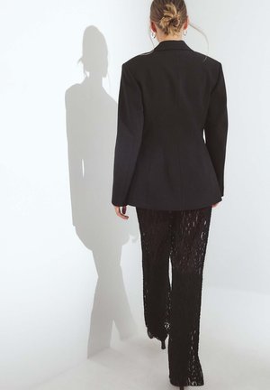 Czarna dopasowana marynarka z ostrymi klapami w zestawieniu z przezroczystymi czarnymi spodniami koronkowymi, tworzącymi warstwowy efekt fakturowy. Cień widoczny na ścianie.