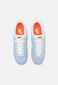 Gaiši zili zamša sporta apavi ar baltām akcentu un šņorēm. Iekšējo odere ir oranža ar Nike logotipu, un tiem ir klasiskā silueta dizains.