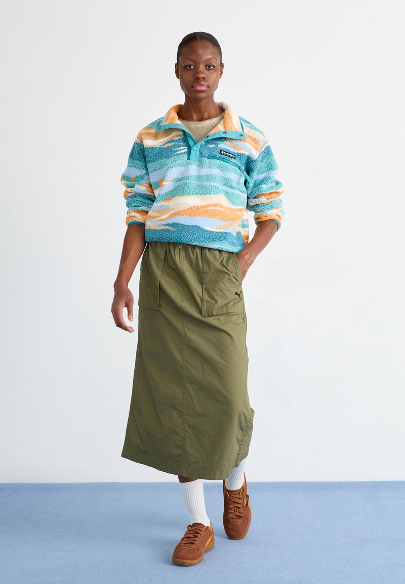 Pull en polaire bleu avec des vagues orange et crème, col montant, associé à une jupe midi verte olive et des baskets brunes sur des chaussettes blanches.