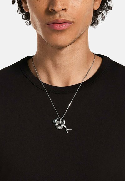 Cou de la personne et partie inférieure du visage portant une chemise noire et un collier en chaîne argentée avec trois charms en pendentif.